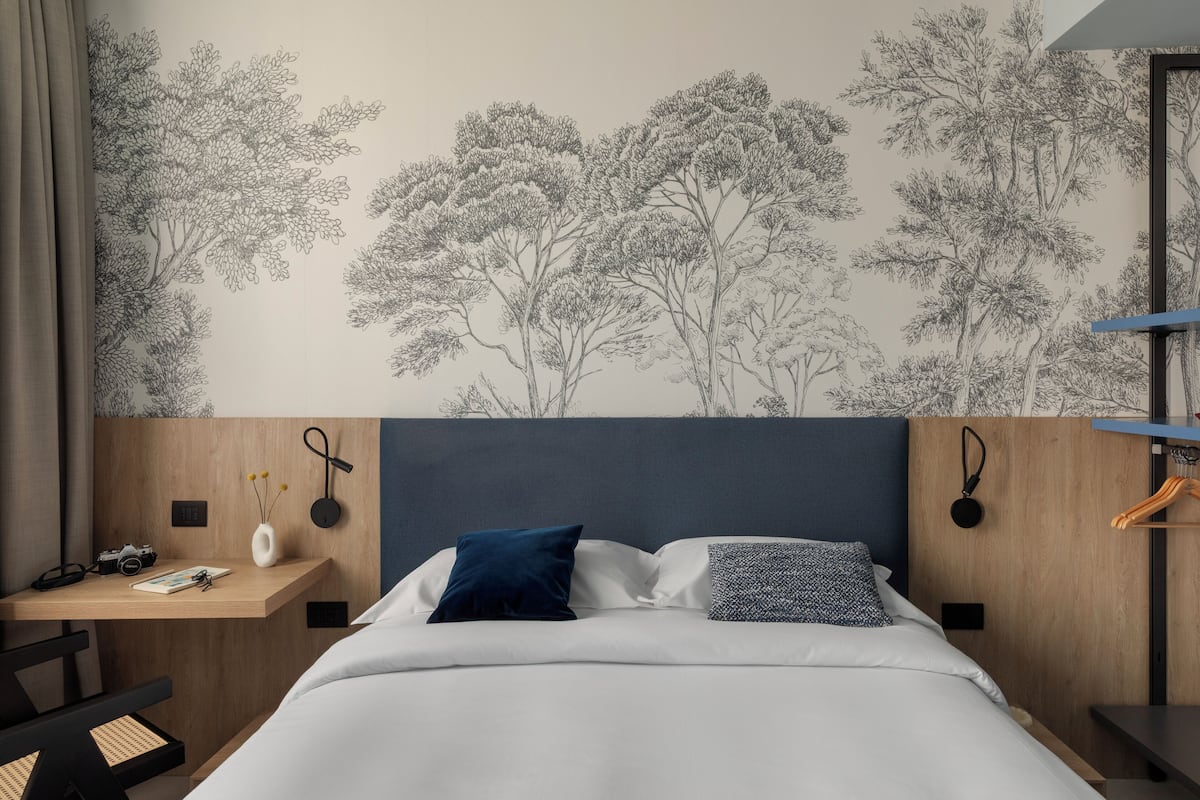 Une chambre d’hôtel avec un lit, un bureau doté d’une caméra, un vase avec des fleurs et un mur orné d’un motif représentant un arbre.