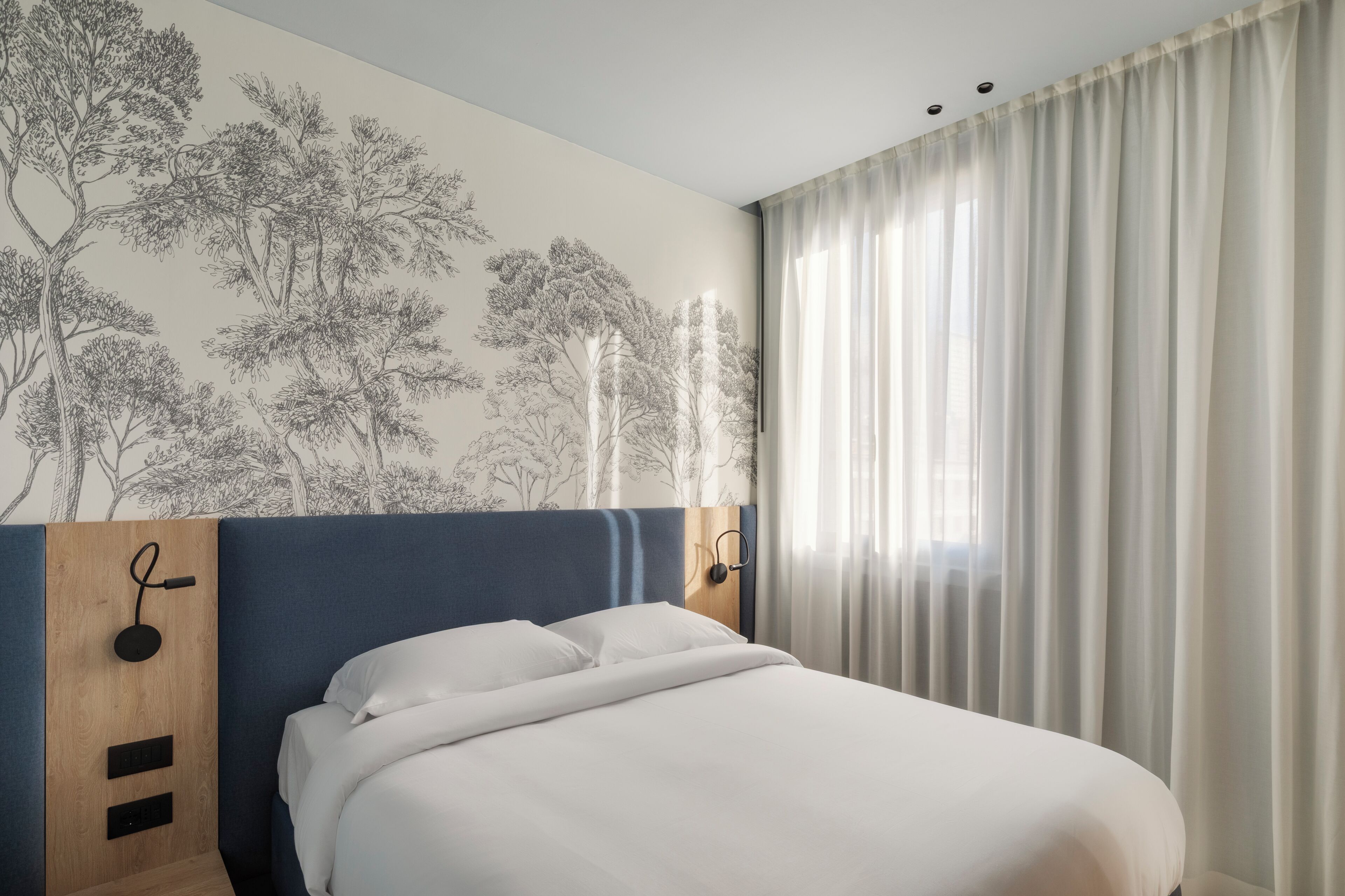 Een opgemaakt bed met witte lakens, een hoofdbord met blauwe accenten, een wandlamp en een raam met dunne gordijnen.