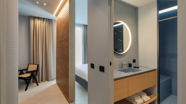 A modern hotel room with a bed, a wooden chair, a bathroom with a sink and mirror, and a shower.