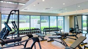 A modern gym with various exercise equipment, large windows, and a view of greenery outside. - Chic and Cozy 1BR Retreat - Canal and City View (Dubai)