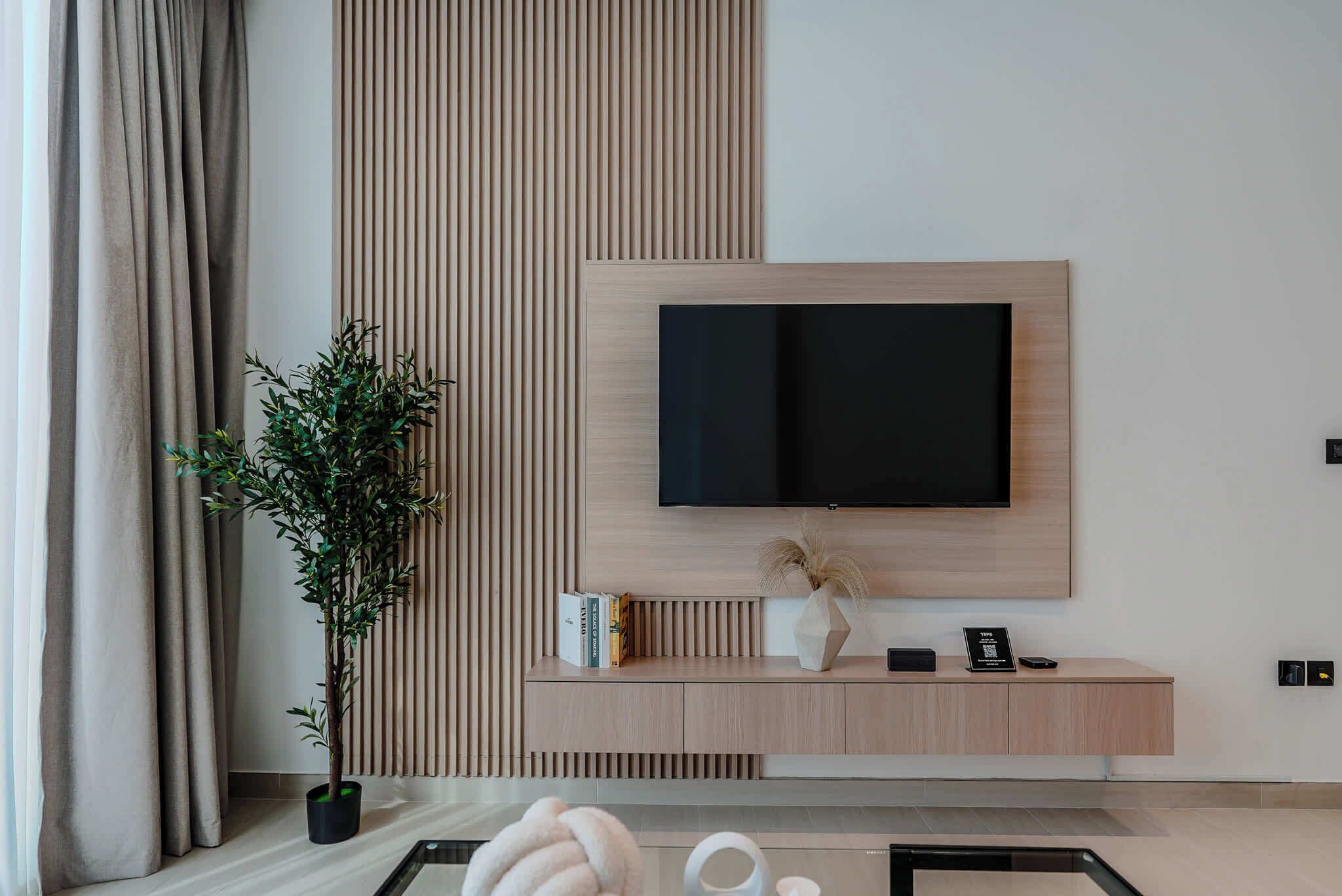 A modern living room with a flat-screen TV mounted on a wooden wall, a potted plant, a glass coffee table, and a minimalist console table with books and decorative items.