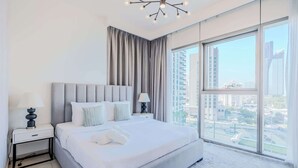 Ein modernes Hotelzimmer mit einem großen Bett, einem Nachttisch, einer Lampe und Blick auf die Stadt durch große Fenster.