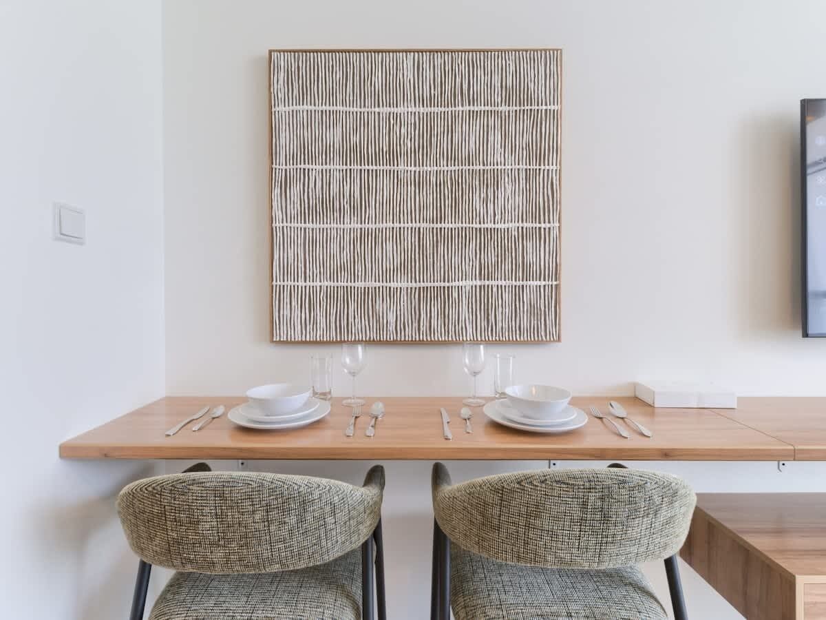 Un comedor con mesa de madera para dos personas, dos sillas de mimbre y un panel decorativo en la pared.