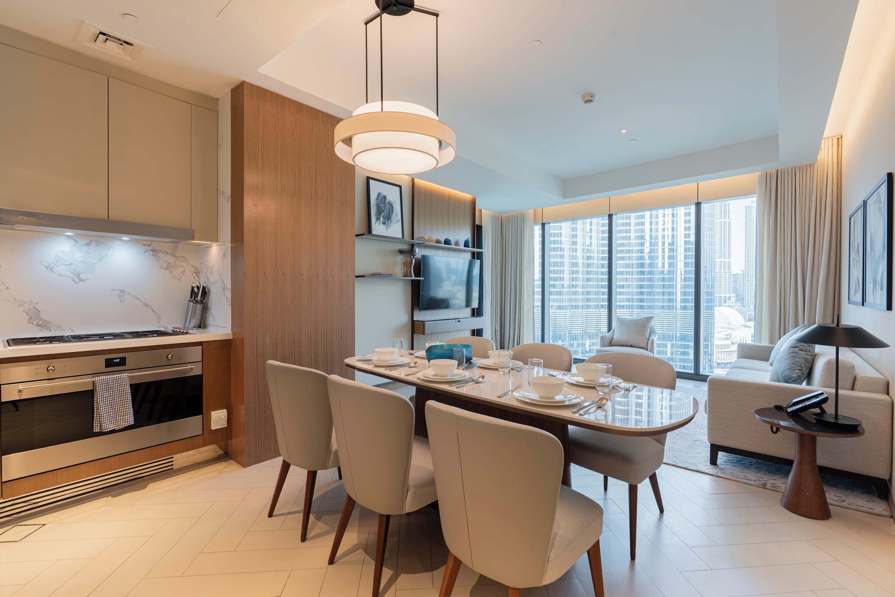 A modern kitchen with a built-in oven, a dining area with a round table and chairs, a living room with a sofa and a TV, and large windows offering a city view.