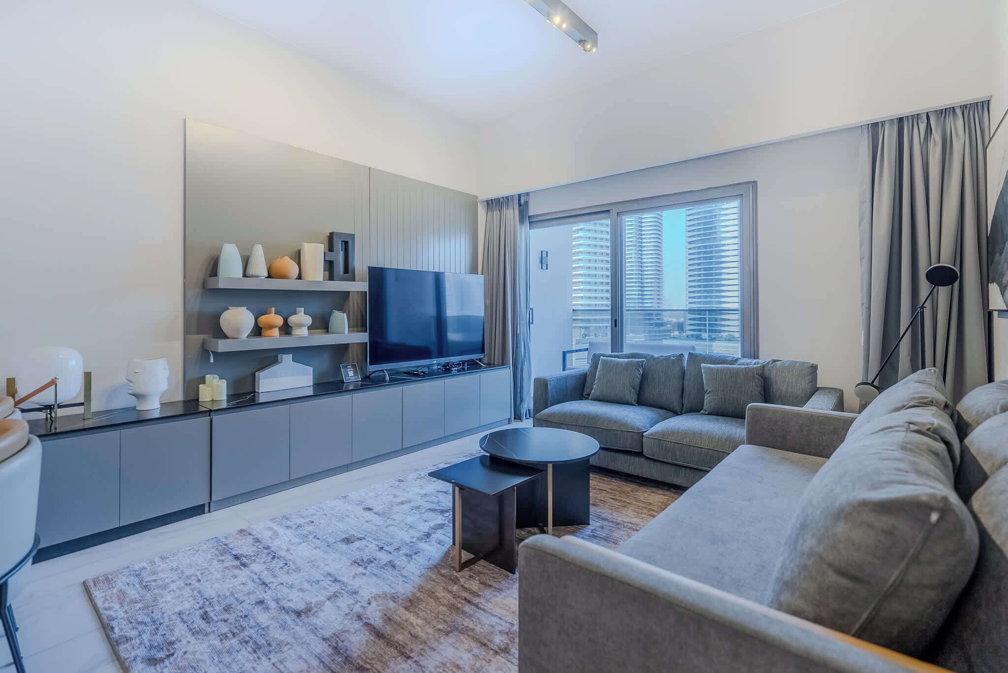 Een moderne woonkamer met een grijze bank, een flatscreen televisie, een salontafel en een groot raam met zonwering.