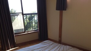 Una habitación con suelo de madera, cama con ropa blanca, ventana con cortinas y una lámpara fijada a la pared.