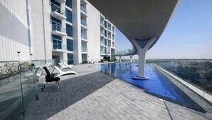 Een dakterras met een zwembad, ligbedden, een glazen balustrade en een modern gebouw met balkons.