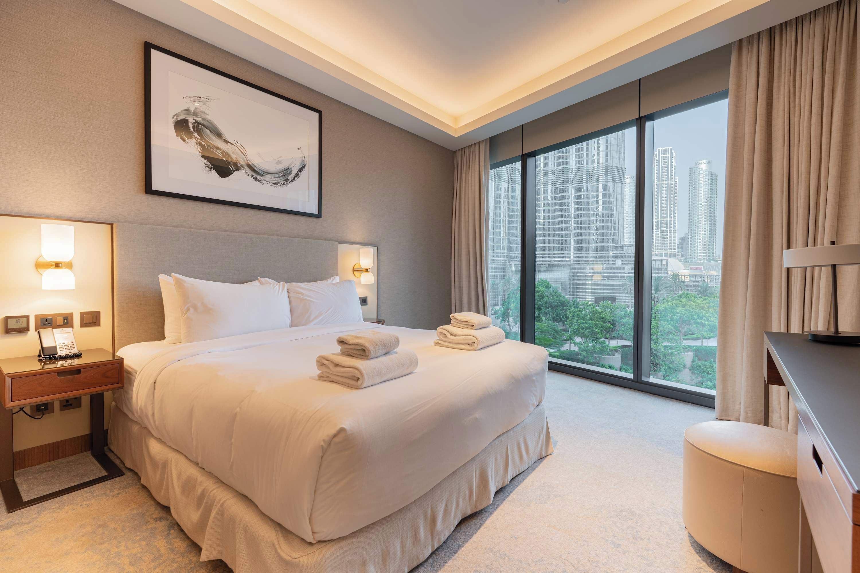 A modern hotel room with a large bed, bedside lamps, a desk, and a view of the cityscape through large windows.