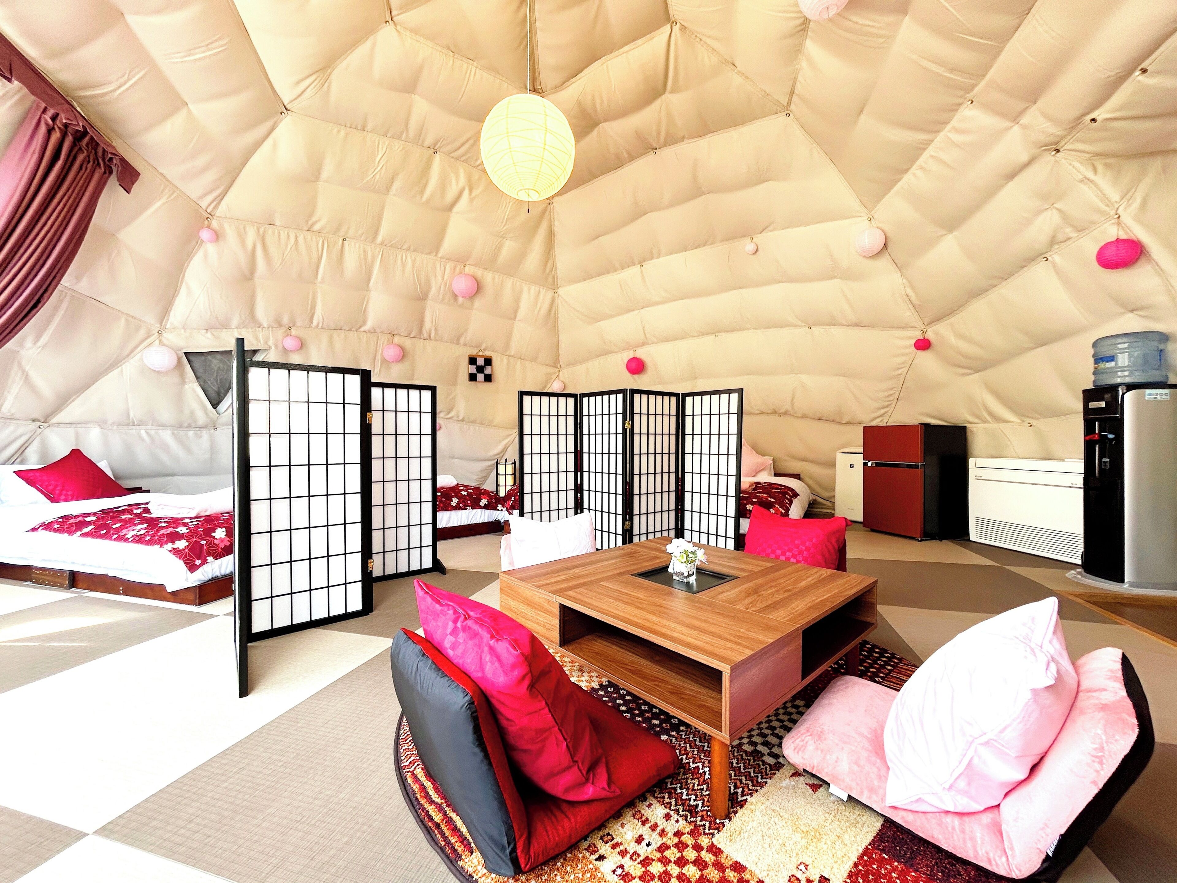Una habitación amplia y luminosa con techo alto, una cama con ropa de cama roja, una mesa de madera y un dispensador de agua.