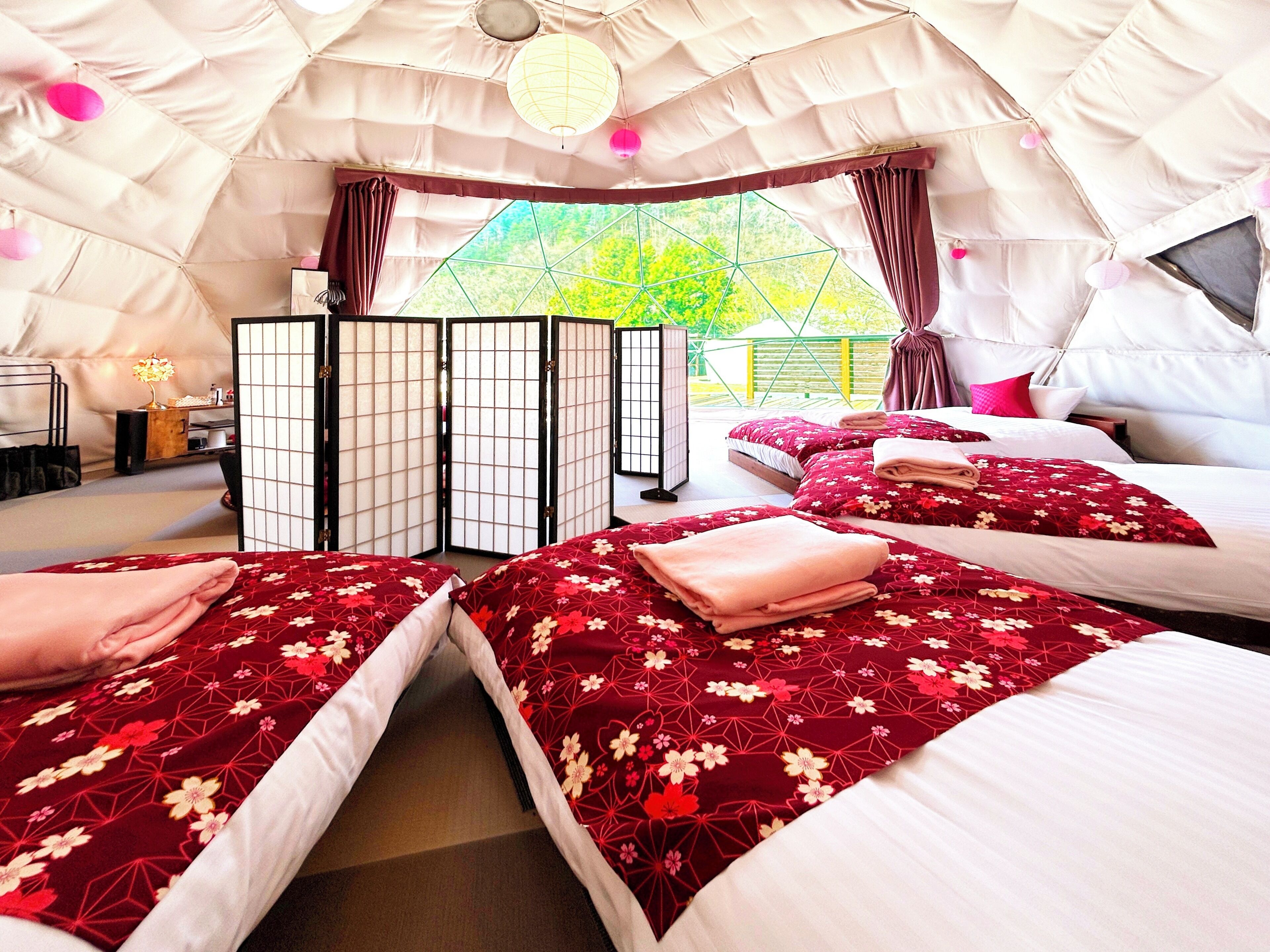 A cozy, well-lit tent-style room with two beds, a traditional Japanese screen, and a view of greenery outside.