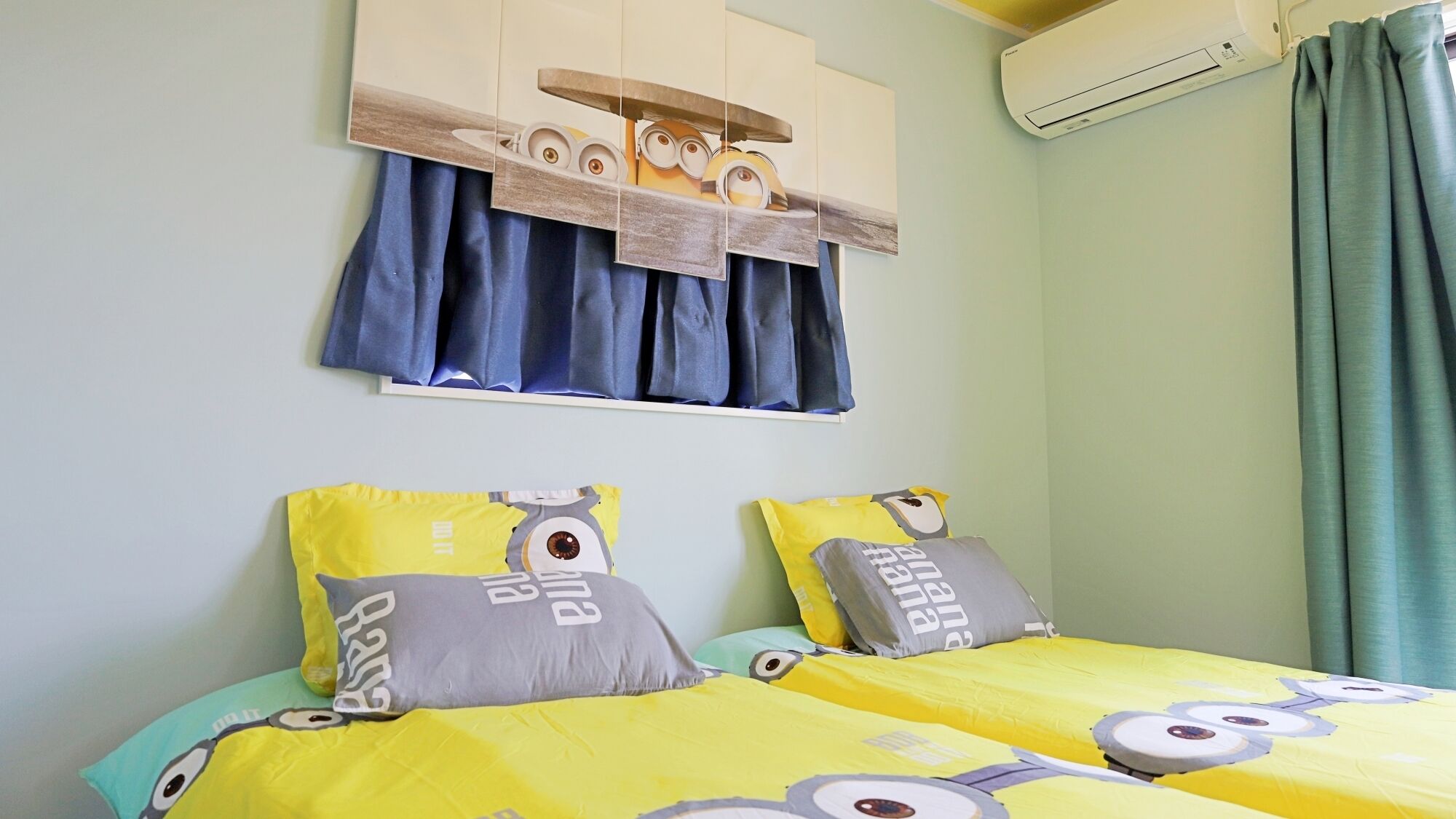 A bedroom with two beds, each with a Minion-themed duvet and pillows, a window with blue curtains, and an air conditioning unit on the wall.