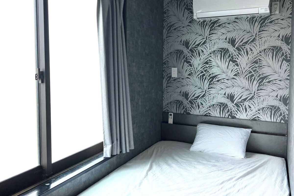 A small, single-bed room with a window, a wall-mounted air conditioner, and a patterned wallpaper.