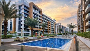 A modern residential building with a rooftop swimming pool, surrounded by palm trees and lounge chairs. - Luxury 2BR Getaway - Prime Location in Meydan (Dubai)