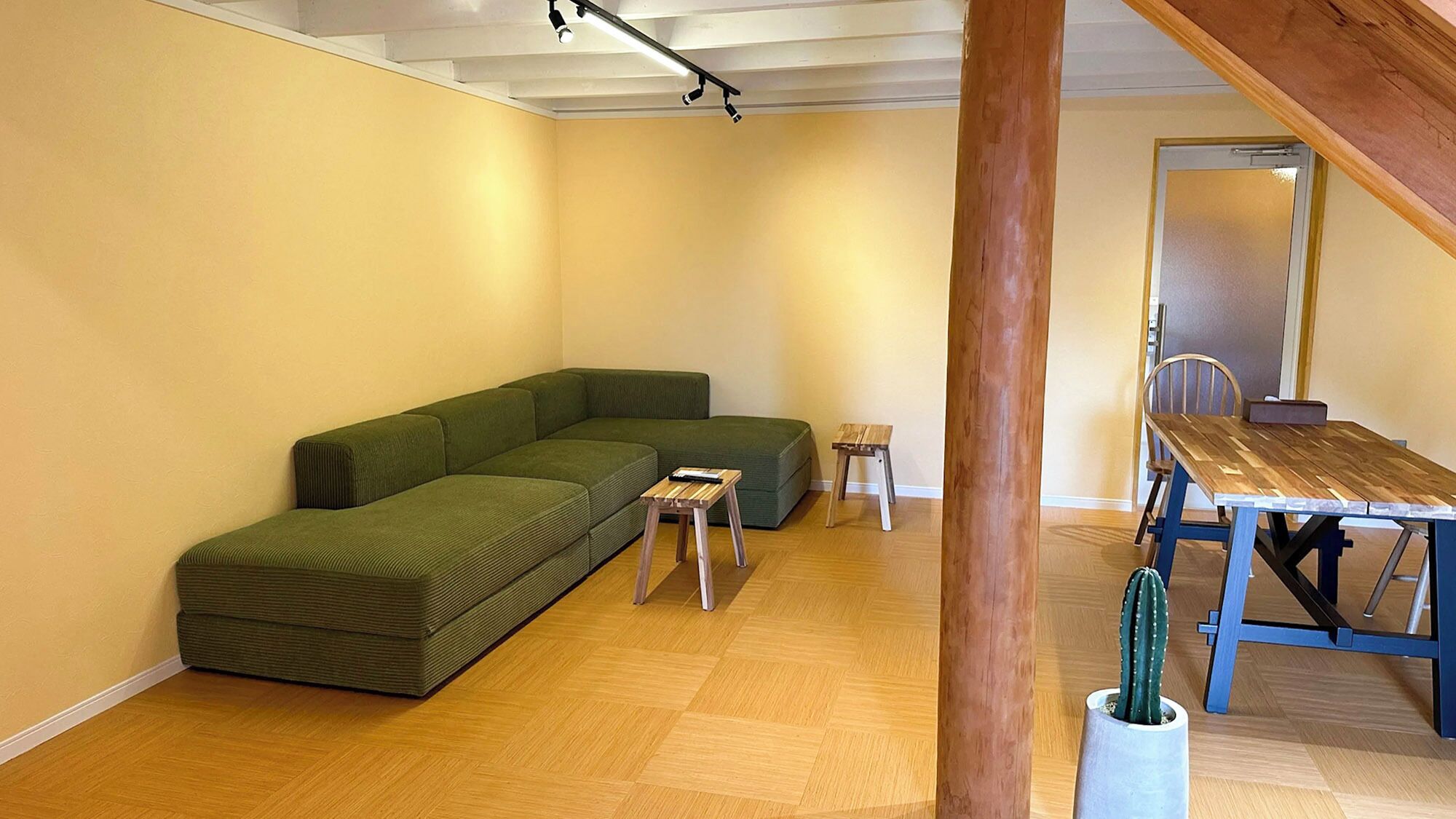 A room with a green sectional sofa, a small wooden table, a wooden dining table, and a cactus plant.