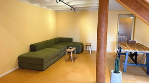 A room with a green sectional sofa, a small wooden table, a wooden dining table, and a cactus plant.