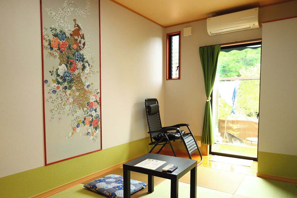 Ein Zimmer mit Tatami-Boden, einem schwarzen Sessel, einem kleinen Tisch mit Fernbedienung und einem großen Blumenbild an der Wand.