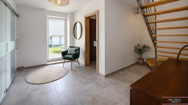 Un intérieur spacieux avec un escalier, un grand miroir rond, un fauteuil vert et une plante en pot.