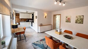 Une cuisine moderne avec des meubles blancs, un coin repas avec une table blanche et des chaises orange, et une vue sur l’espace de vie adjacent.