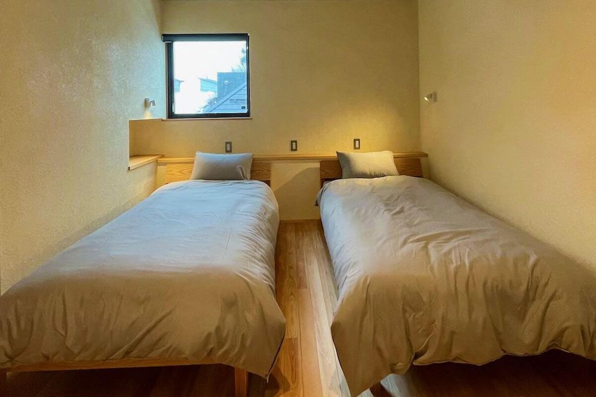 Une petite chambre bien éclairée, avec deux lits simples, une fenêtre et une table de chevet avec une lampe.