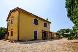 A two-story yellow building with a tiled roof, a gravel driveway, and a covered porch area. - Le Torrette Etrusche Room 6 – Comfort and Relaxation in Capalbio (Capalbio)