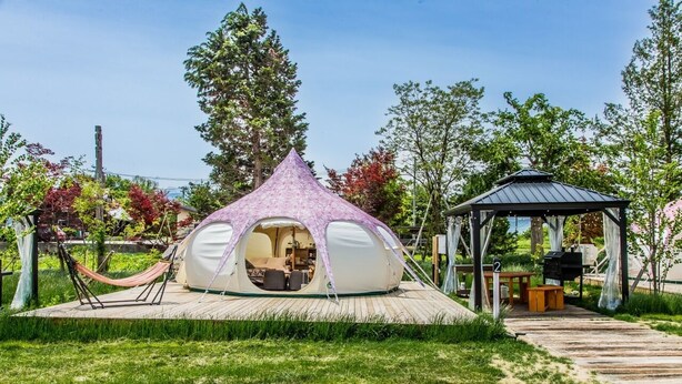 Estructura de cúpula geodésica con interior abierto, terraza de madera y un pabellón en un jardín ajardinado.