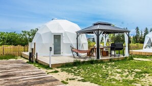 Una estructura de domo geodésico blanco con terraza de madera, una hamaca, una parrilla y un pabellón.