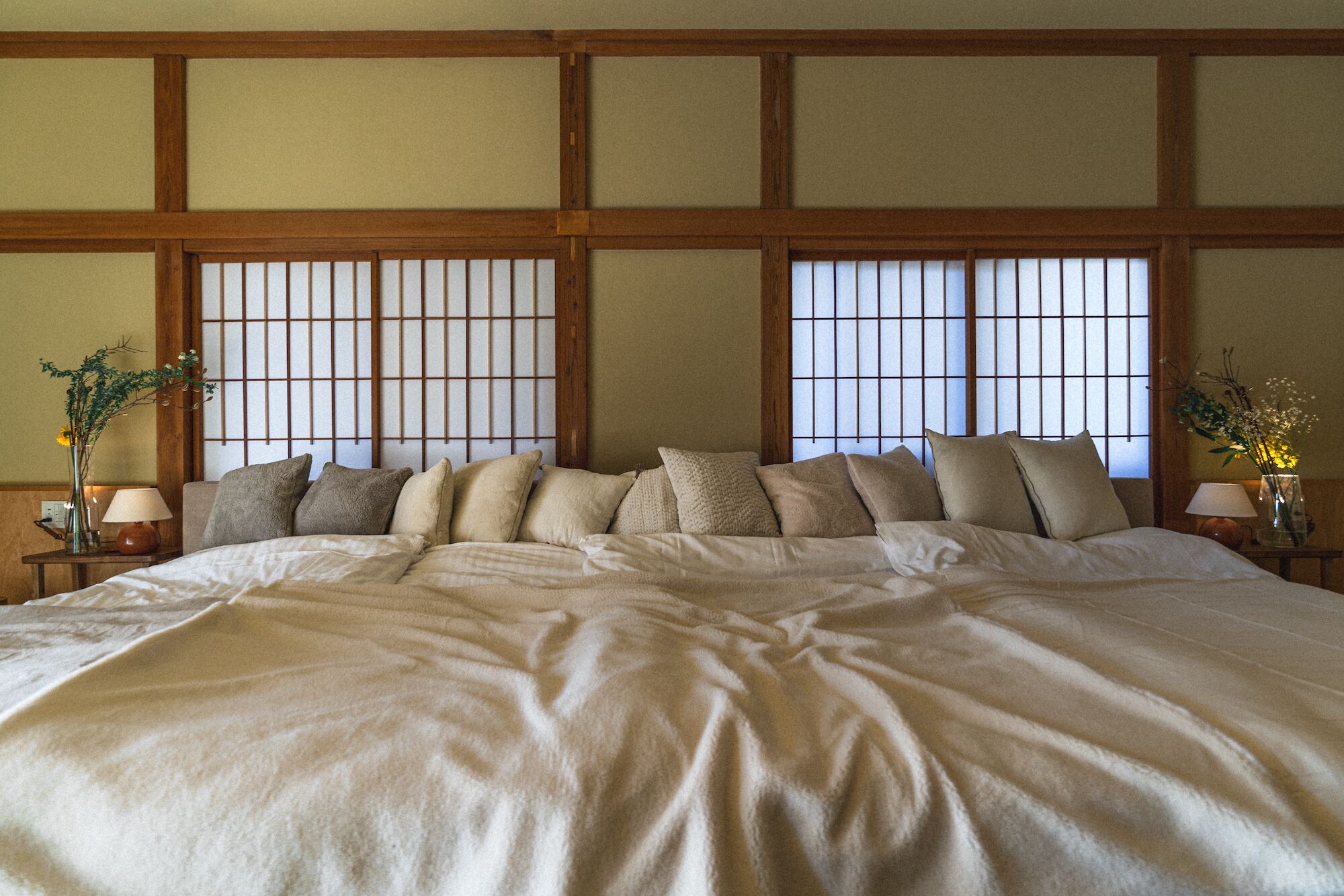 Una cama grande con varias almohadas, cabecera de madera y una ventana tradicional al estilo japonés con diseño de cuadrícula.