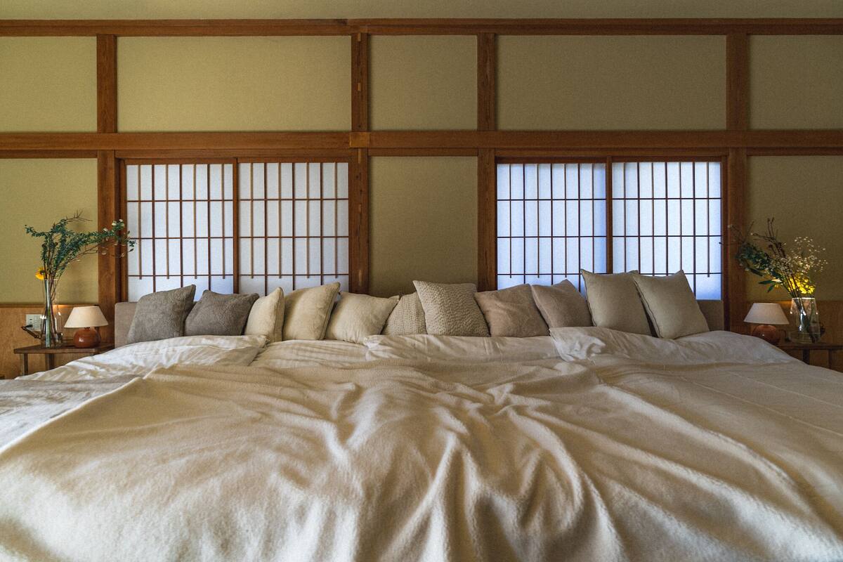 A large bed with multiple pillows, a wooden headboard, and a traditional Japanese-style window with a grid pattern.