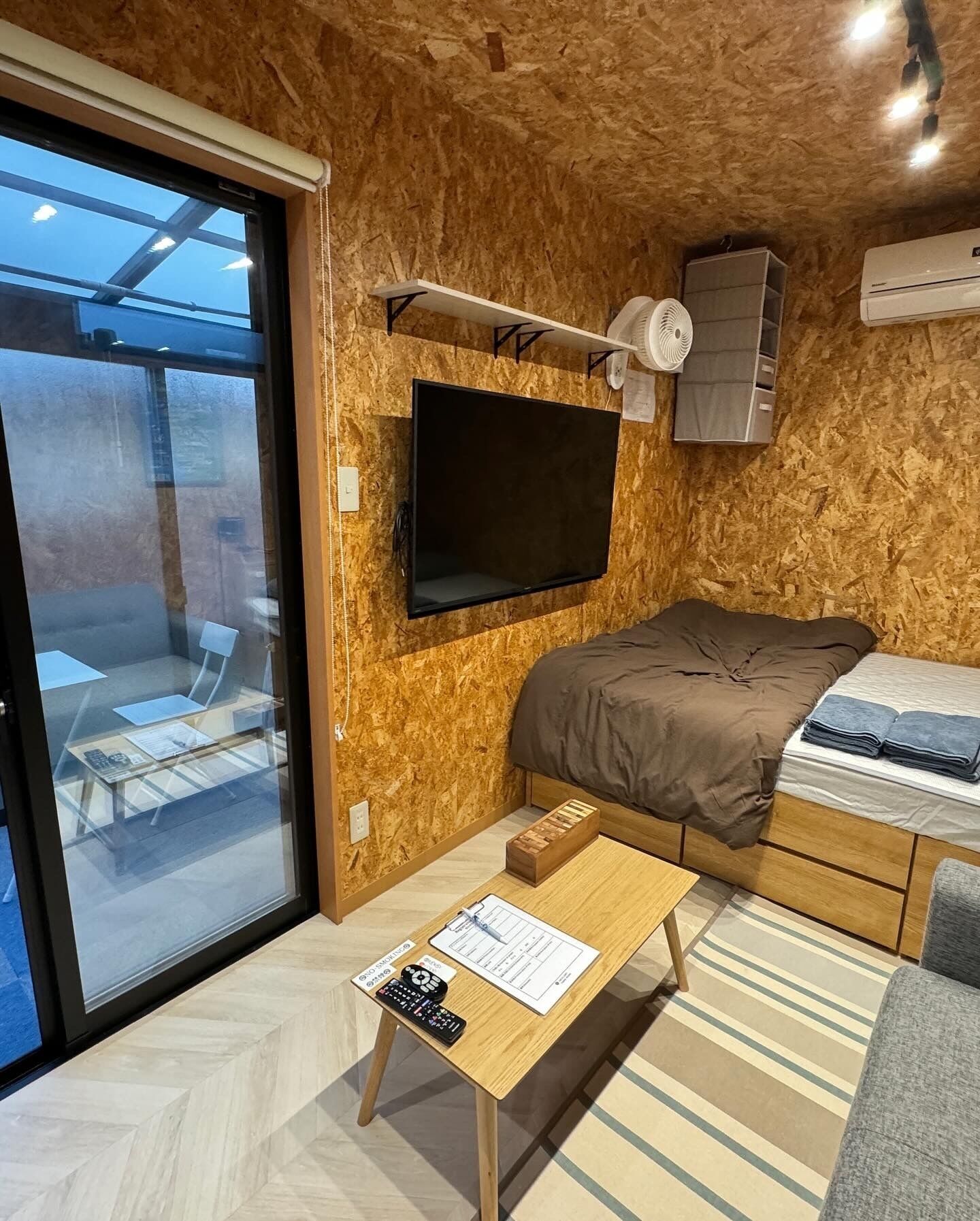 A compact room with a bed, a mounted TV, a small table with a remote, and a window with a view of outdoor seating.