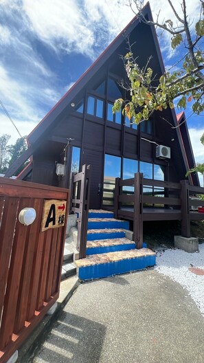 Un edificio con techo inclinado, una puerta de madera con un letrero con la letra 'A' y una escalera que lleva a la entrada.