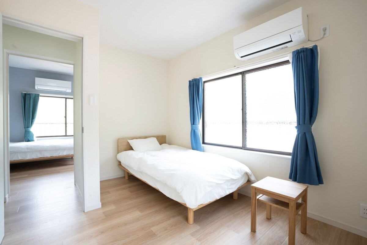 A bedroom with a bed, a bedside table, a window with curtains, an air conditioning unit, and a hallway leading to another room.