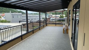 A covered balcony with a metal railing, a bench, and a view of buildings and greenery. - Irorian (Kamakura)