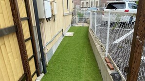 A narrow, enclosed outdoor area with artificial green turf, a chain-link fence, and a building with beige siding and a window. - Irorian (Kamakura)