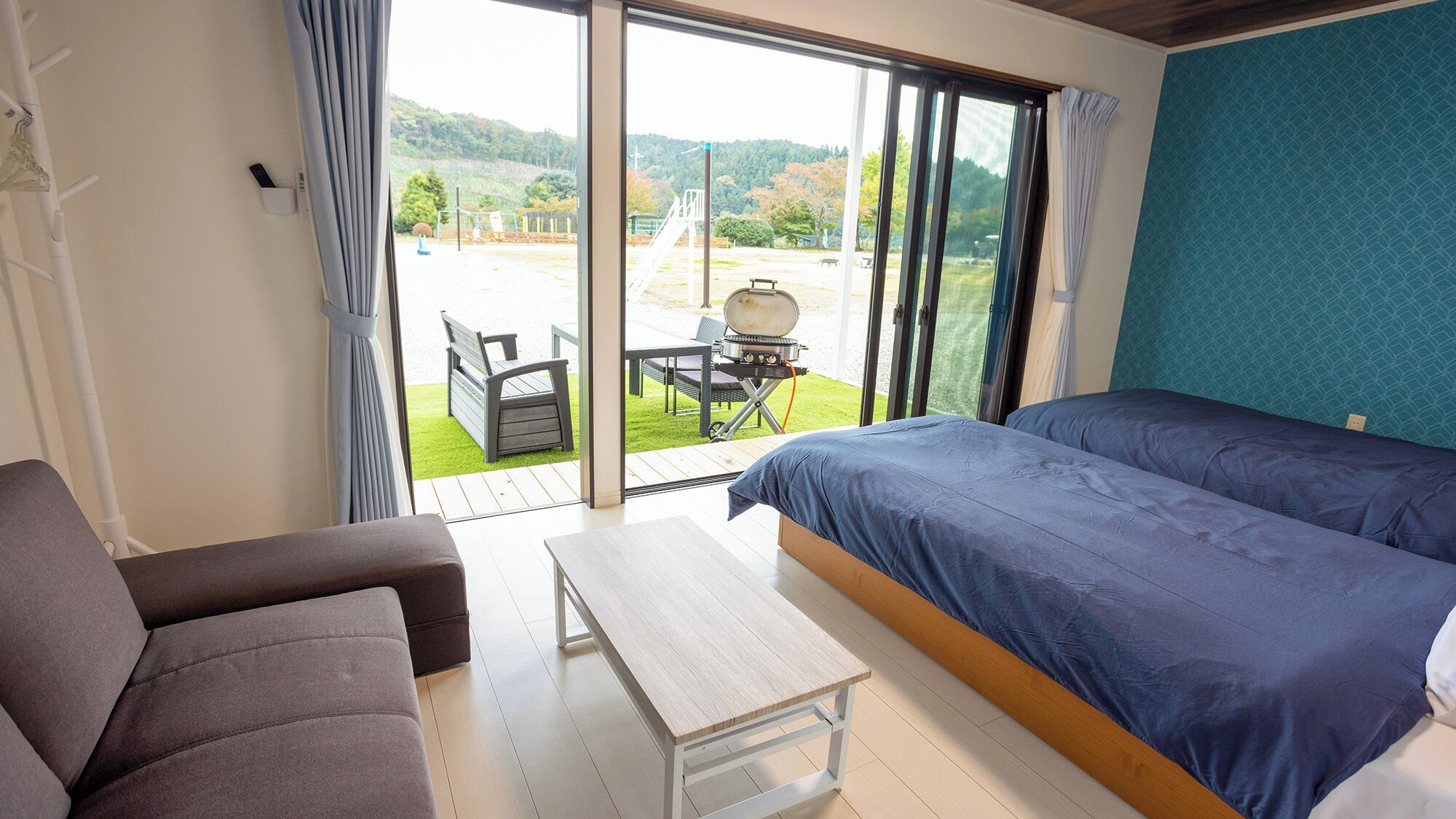 A room with a bed, a sofa, a small table, and a view of a grassy area with a barbecue and a playground.