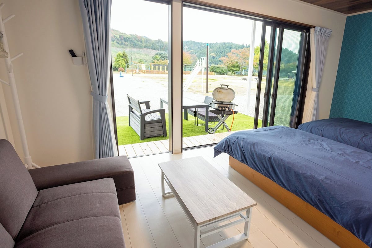 A room with a bed, a sofa, a small table, and a view of a grassy area with a barbecue and a playground.