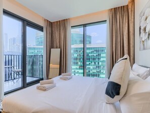 Ein Hotelzimmer mit einem großen Bett, zwei Vorhängen, einem Balkon mit Tisch und Stuhl und Blick auf eine Stadtlandschaft.