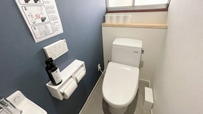 A small bathroom with a toilet, a sink, and a paper towel dispenser. There is a window above the toilet and a shelf with toilet paper. - Marine Amkusa 2gokyo (Takaharu)