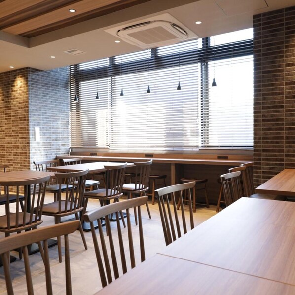 Un interior de restaurante con mesas y sillas de madera, amplias ventanas con persianas e iluminación colgante.