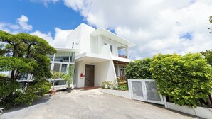 A modern white house with a large balcony, surrounded by lush greenery and a gated entrance.