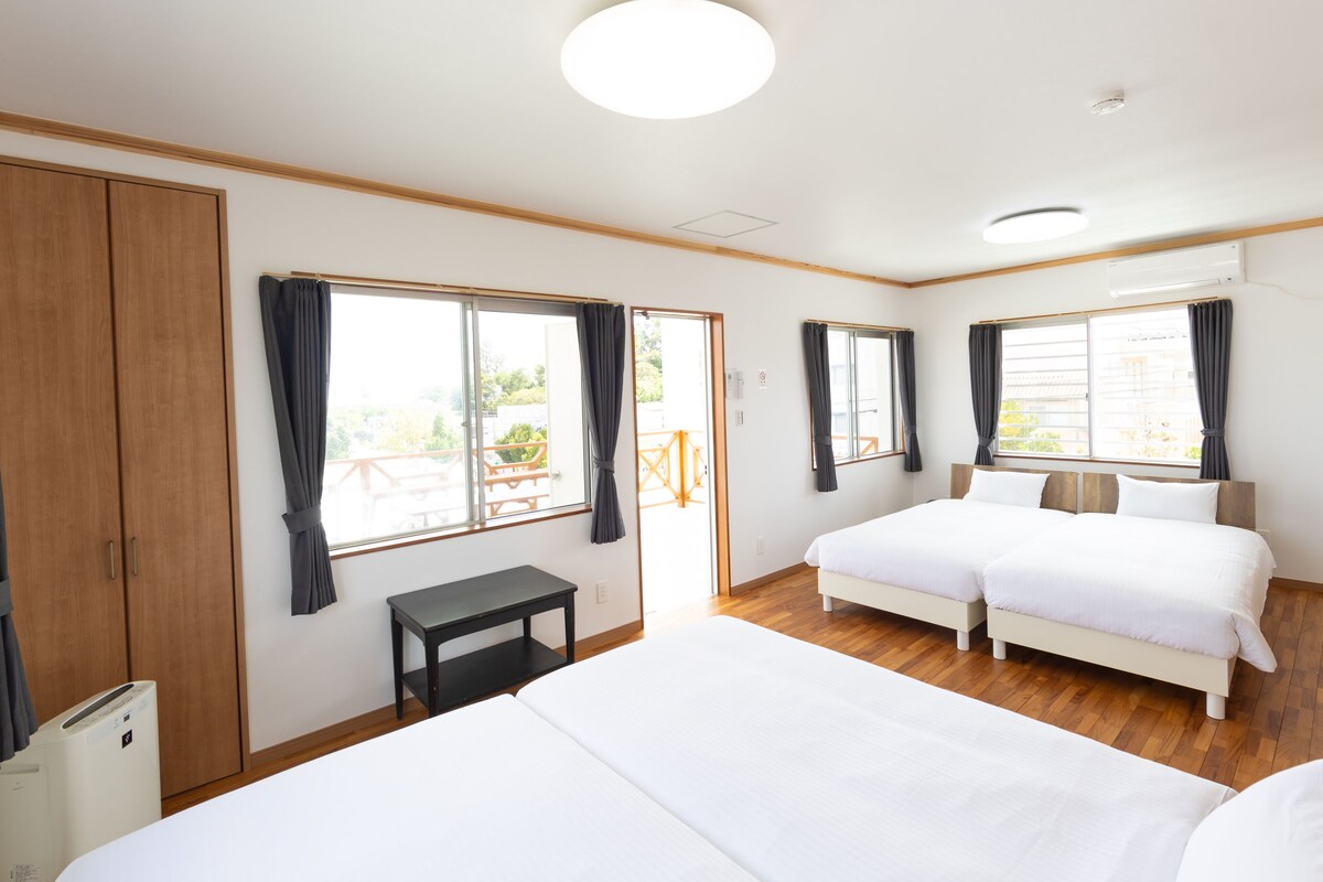 Une chambre d’hôtel avec deux lits, une petite table et une armoire. De grandes fenêtres sont présentes, avec des rideaux, et on peut voir un appareil de climatisation fixé au mur.