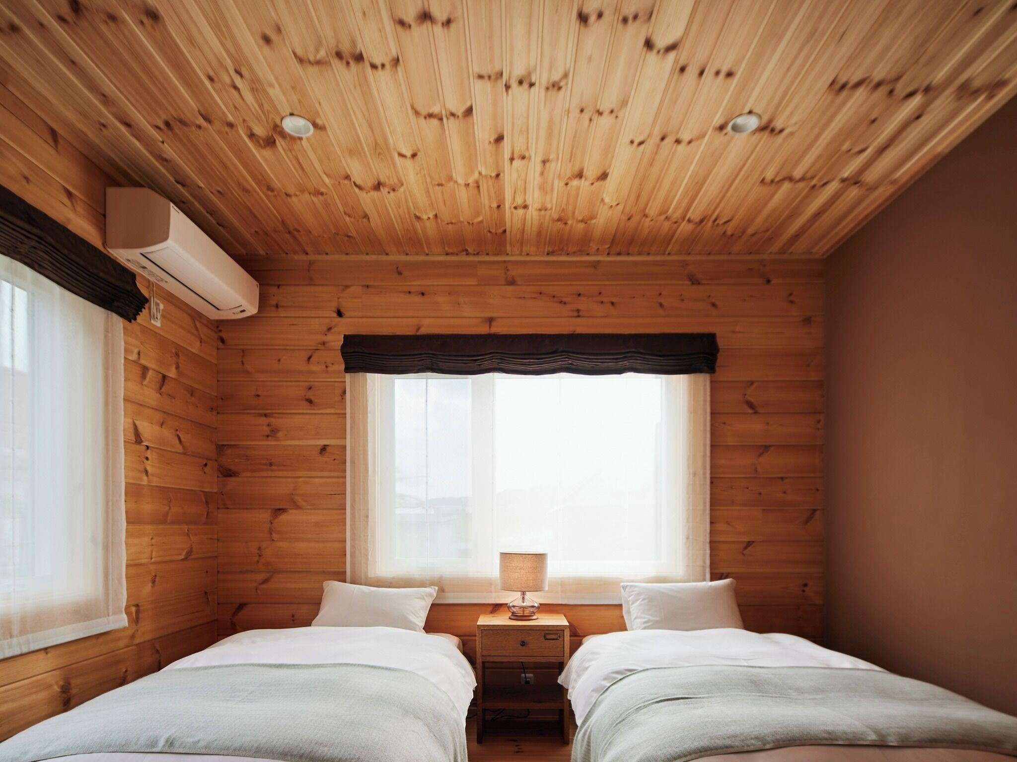 A cabin-style bedroom with two beds, a window with a dark valance, and a nightstand with a lamp.