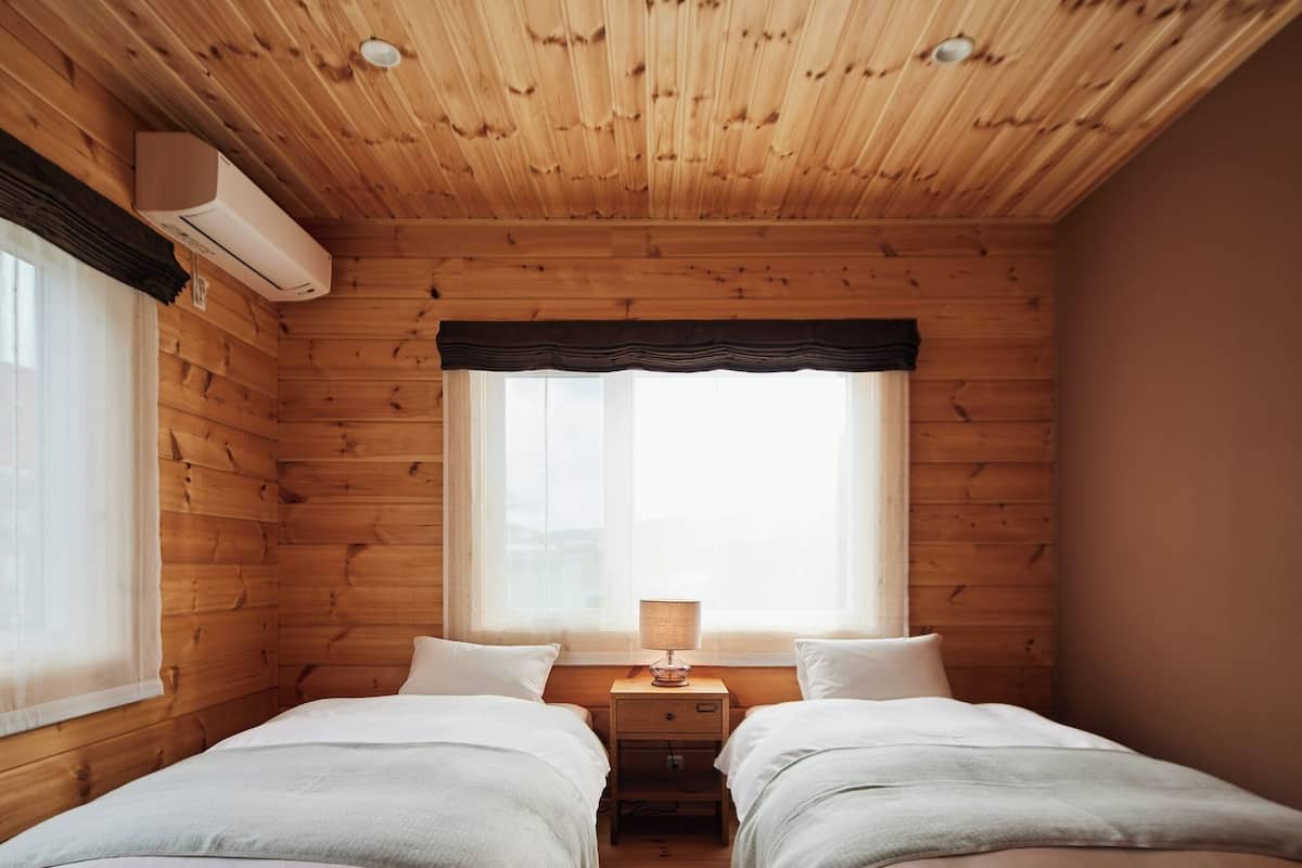 Un dormitorio tipo cabaña con dos camas, una ventana con cortina oscura y una mesita de noche con lámpara.