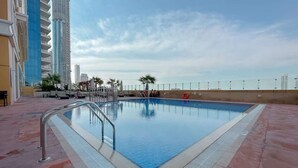 Ein Schwimmbecken mit Metallleiter, umgeben von einer gefliesten Terrasse, mit Hochhäusern im Hintergrund.