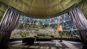 Una habitación con tienda de campaña y vistas a un bosque, un sofá verde, una mesa de madera y una lámpara.