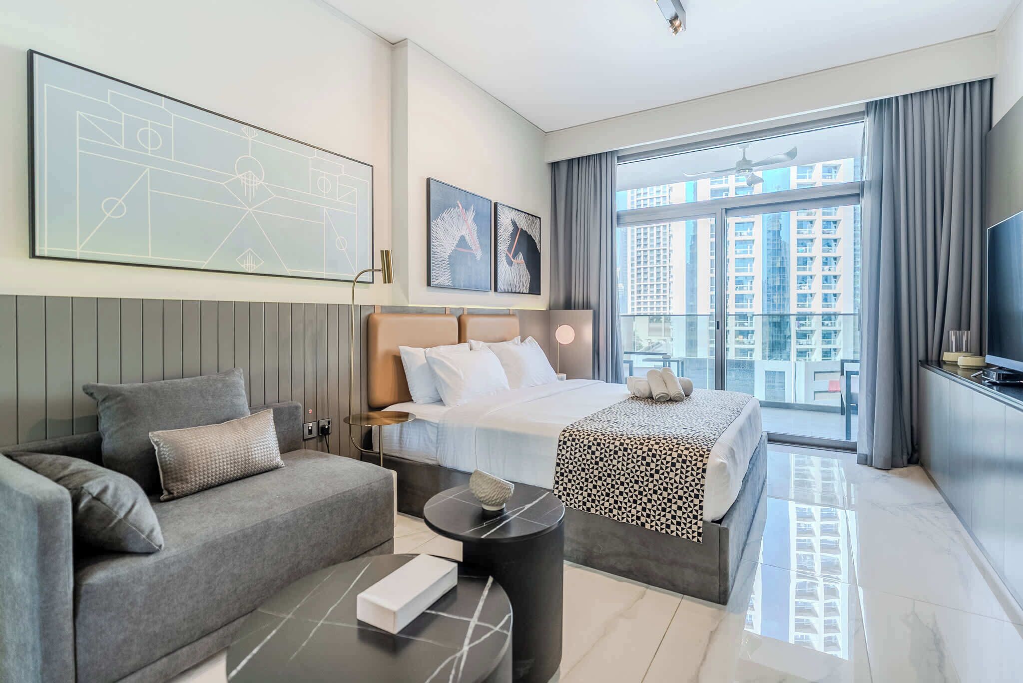 A modern hotel room with a bed, a sofa, a small table, and a large window offering a city view.