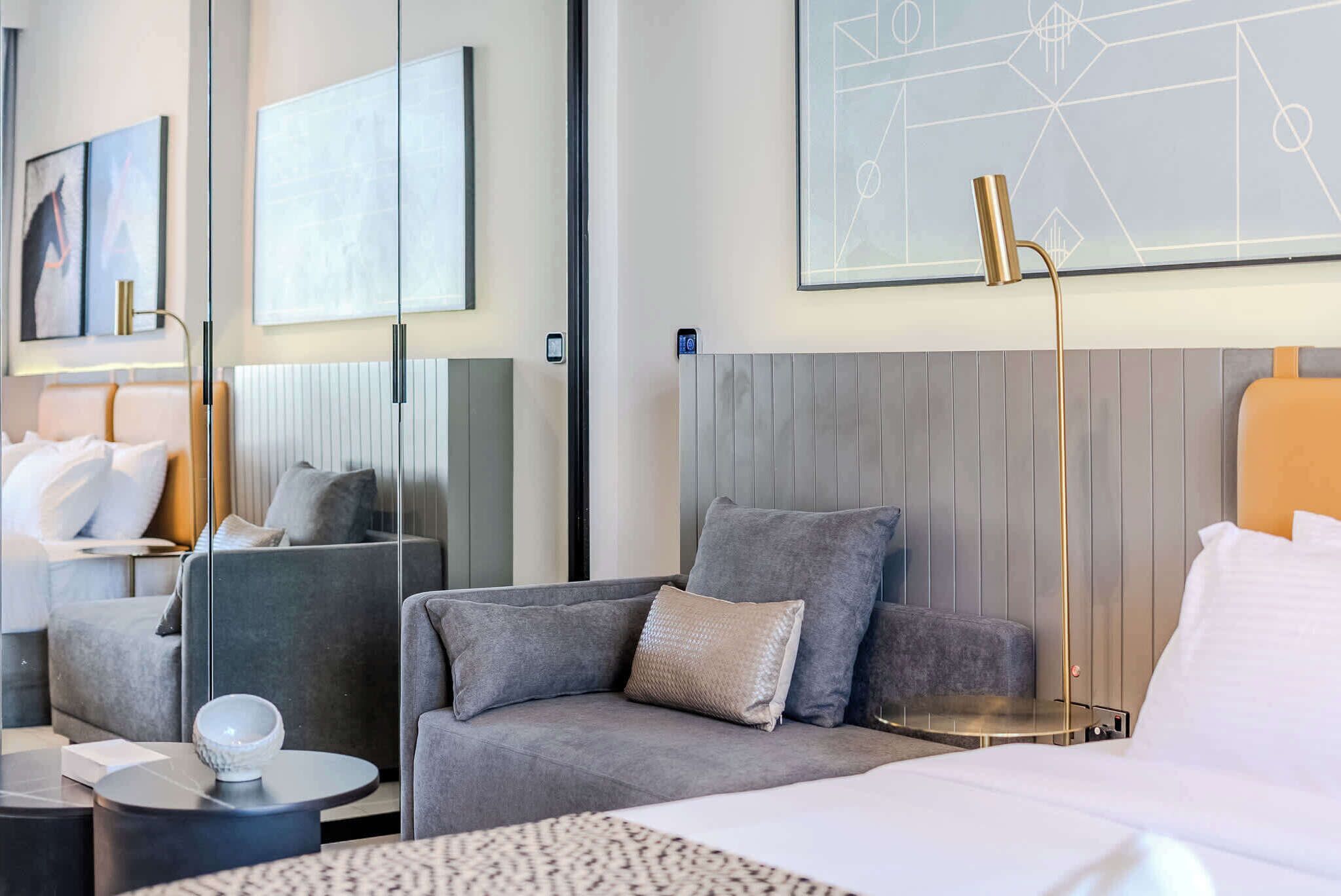 Une chambre d’hôtel moderne avec un canapé gris, une petite table ronde, un lit avec du linge de maison blanc et un grand tableau abstrait accroché au mur.