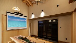 A room with a wooden table, chairs, a projector screen displaying a beach scene, and a sliding door. - B/C Hotel Naha Rent-a-car Hotel (Ishigaki)