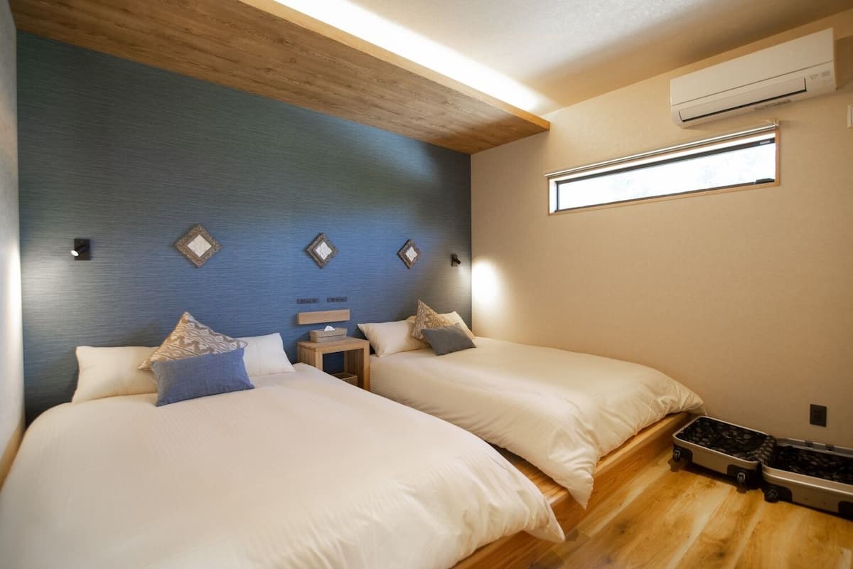 A hotel room with two beds, a wooden ceiling, an air conditioning unit, and a window.