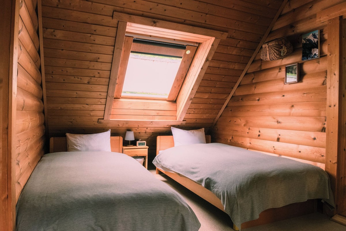 Eine Holzhütte mit zwei Betten, einem kleinen Tisch, einem Nachttisch, einer Uhr und einem Fenster mit Aussicht.