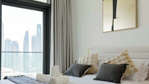 Une chambre d’hôtel avec une grande fenêtre, un lit avec une literie blanche, des oreillers gris et blancs, et une vue sur de hauts immeubles.